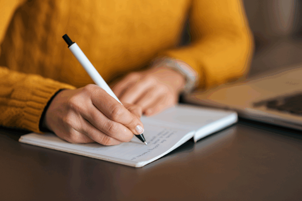 person in orange sweater writing with a pen on a notebook at a desk